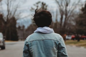 Un jeune, de dos avec une veste en jean, marchant sur une route.
