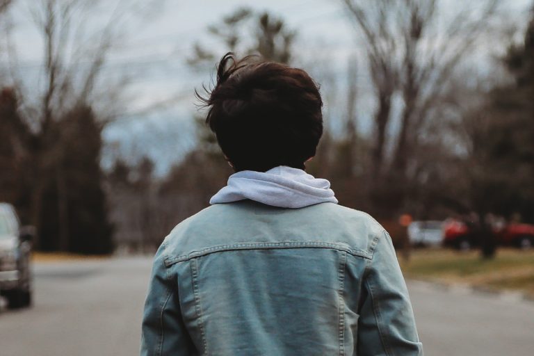 Un jeune, de dos avec une veste en jean, marchant sur une route.